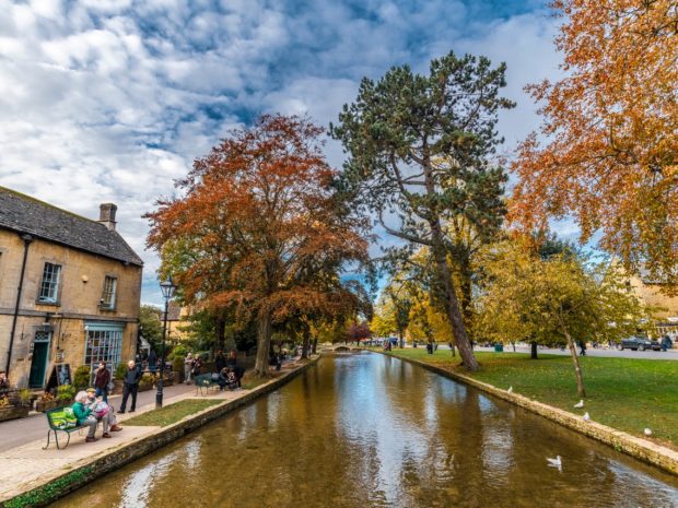 Autumn landscape view of Bourton on the Water in the Cotswolds with trees, river, blue sky, clouds and traditional stone building on the left.