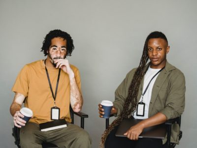 Two young Black men sitting on black chairs wearing casual clothing. One has laptop.