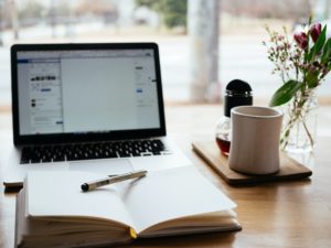 An open notebook lies open on a desk next to a white mug and an open laptop.