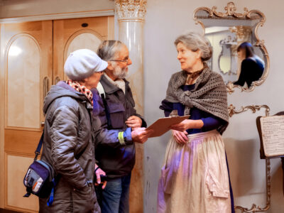 Volunteer in historical dress speaking to two elderly visitors.