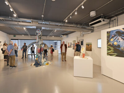 A white art gallery with a number of people viewing art near the back of the room. There are prints on the walls and sculptures made of material on plinths. A column of customised denim stands in the middle of the room.