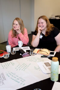 Two white women in their 30s with long blonde hair are smiling and sat at a table with pages of written notes on it.
