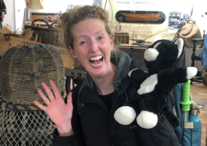 A white woman with curly blonde hair is smiling with both hands up in excitement. One hand has a cat puppet on it.
